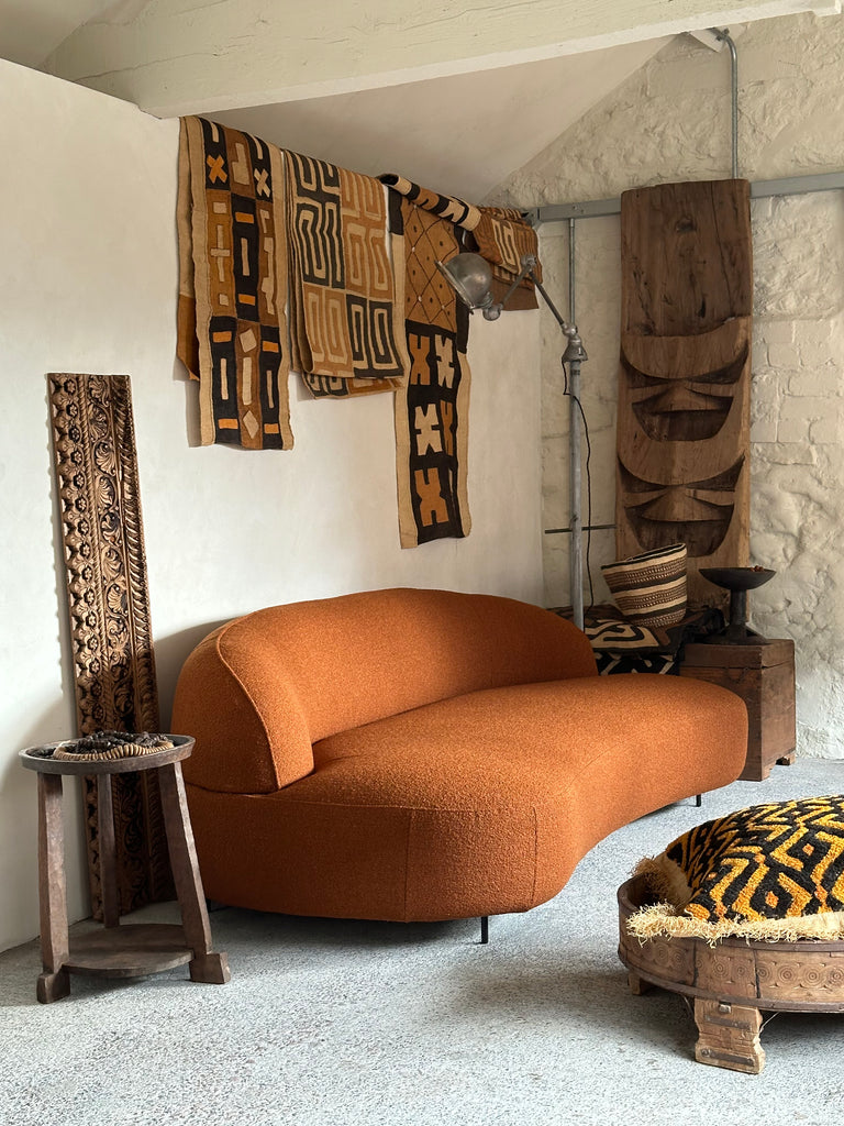 the alia two seater sofa in burnt orange boucle fabric sits in the centre of the image. it has soft curves, a low back and discrete black metal legs. it is surrounded by kuba cloth panels and cushions and intricate pieces of carved wood