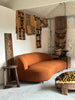 the alia two seater sofa in burnt orange boucle fabric sits in the centre of the image. it has soft curves, a low back and discrete black metal legs. it is surrounded by kuba cloth panels and cushions and intricate pieces of carved wood