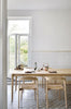 A modern oak dining table with chairs around it, placed in a kitchen setting with a window and a rustic wall shelf in the background.