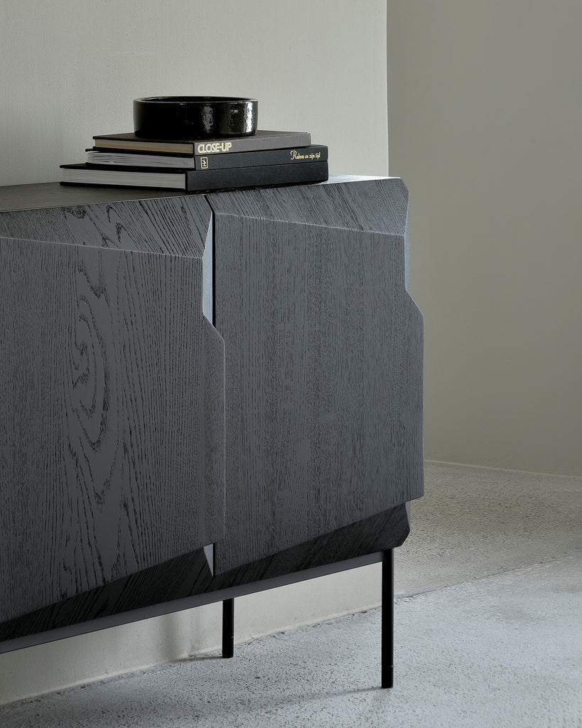 a cropped lifestyle photograph of a black sideboard at a diagonal angle. the doors of the sideboard are roughly square and have a 3d geometric shape to them which includes the handles. it has thin black cylindrical metal legs and a stack of black coffee table books sitting on top.
