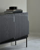 a cropped lifestyle photograph of a black sideboard at a diagonal angle. the doors of the sideboard are roughly square and have a 3d geometric shape to them which includes the handles. it has thin black cylindrical metal legs and a stack of black coffee table books sitting on top.